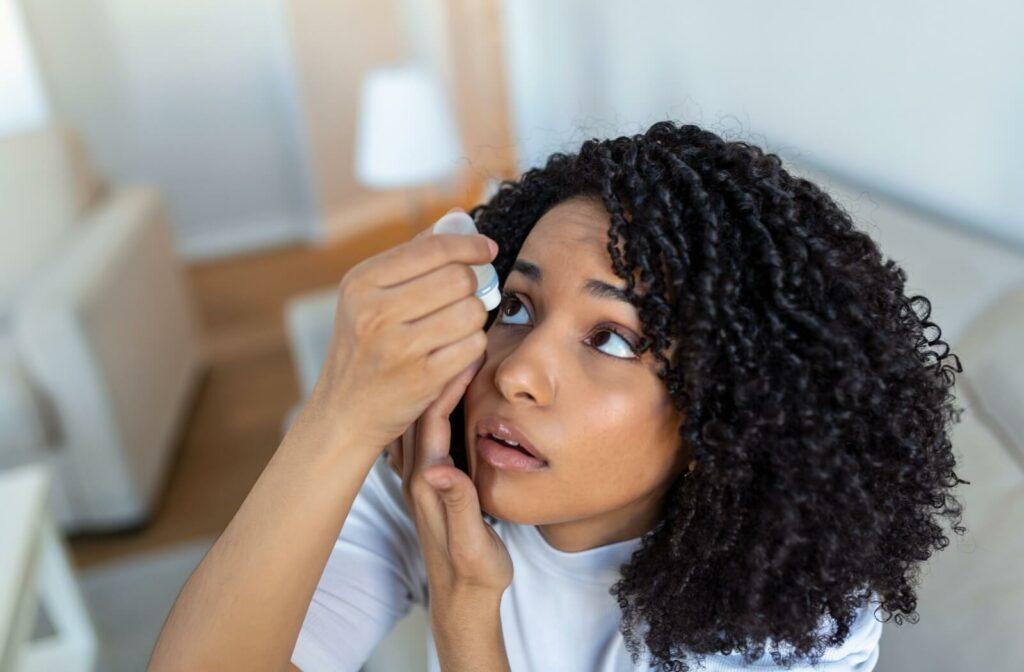 A young person sitting indoors tilting their head back and carefully applying eye drops to relieve dry eyes