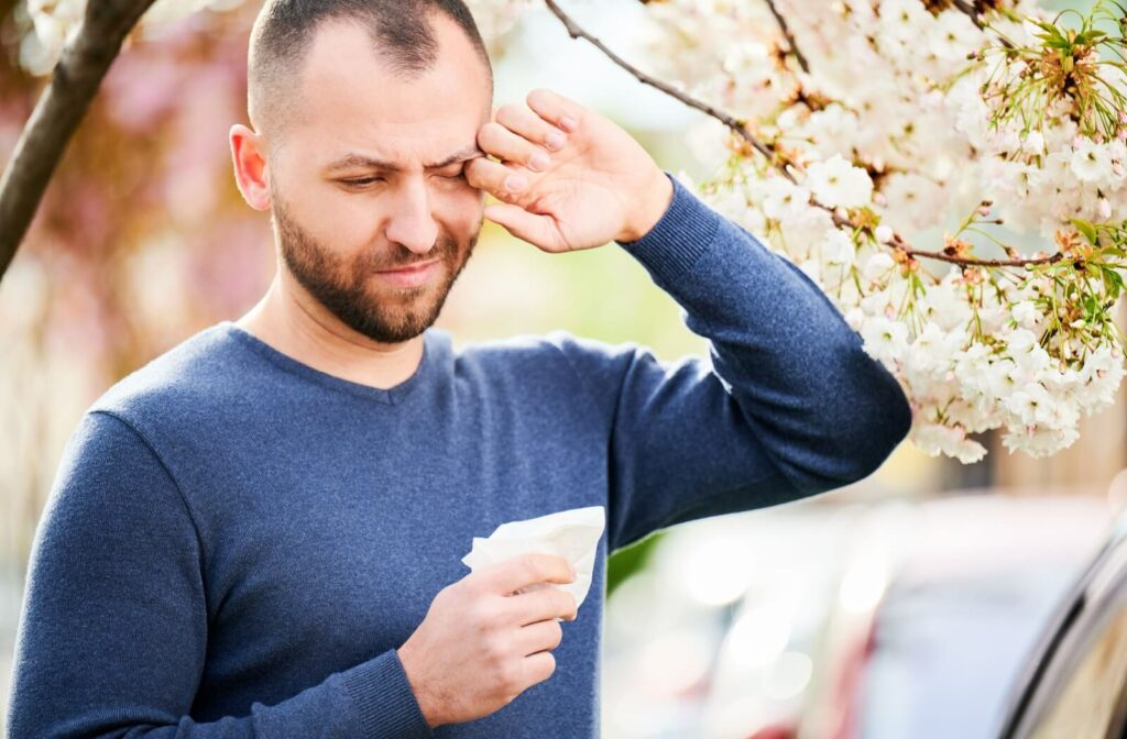 A person standing outside near a blooming tree itching their eye with their hand and a tissue in their other hand