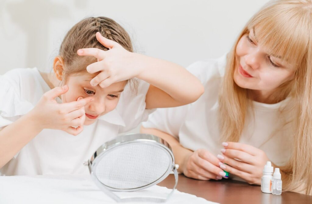 Child putting on contact lenses in front of a desktop mirror while adult seated next to them looks on.