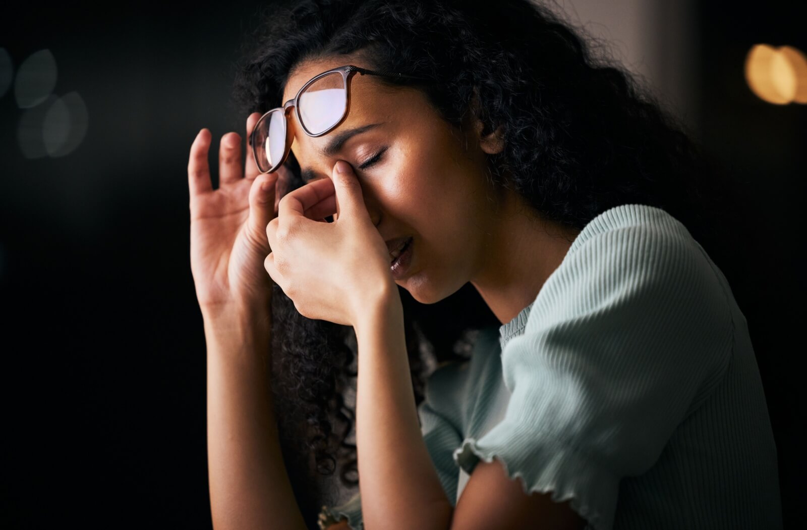 Person with curly hair lifting their glasses and rubbing the bridge of their nose, appearing to experience eye strain or discomfort in a dimly lit setting.