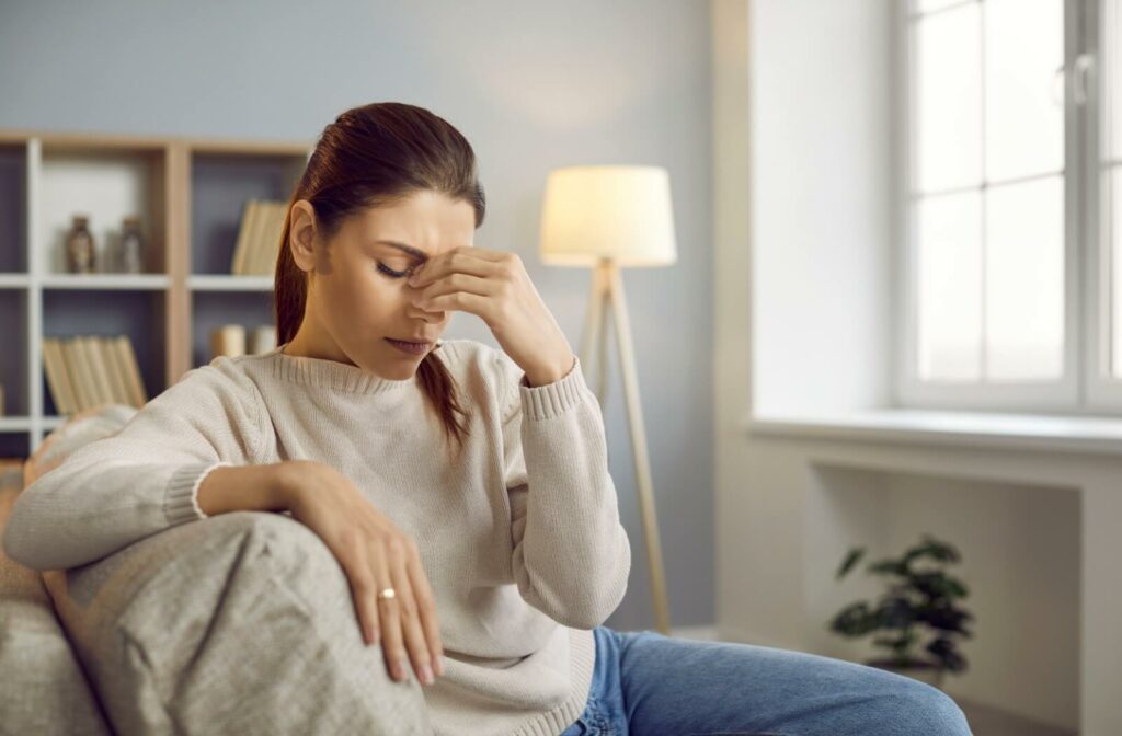 A person sitting on a sofa pinching the bridge of the nose with the eyes closed, illustrating the symptoms of eye fatigue and dizziness caused by visual strain.