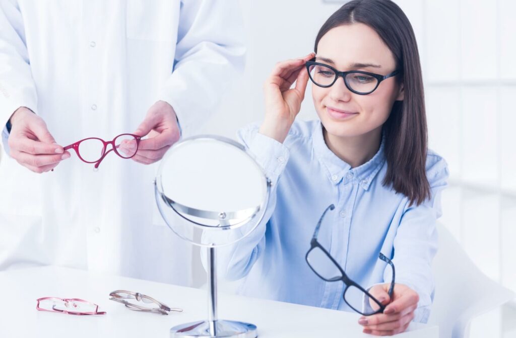 A person trying on black-rimmed glasses while looking in a mirror at an optical shop, with an optometrist holding another pair of red frames nearby.